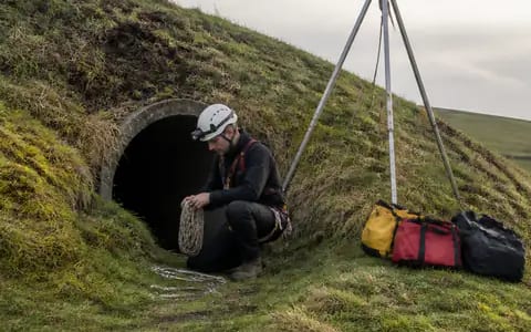 A rescuer in a helmet kneels at a culvert entrance, holding rope beside a tripod